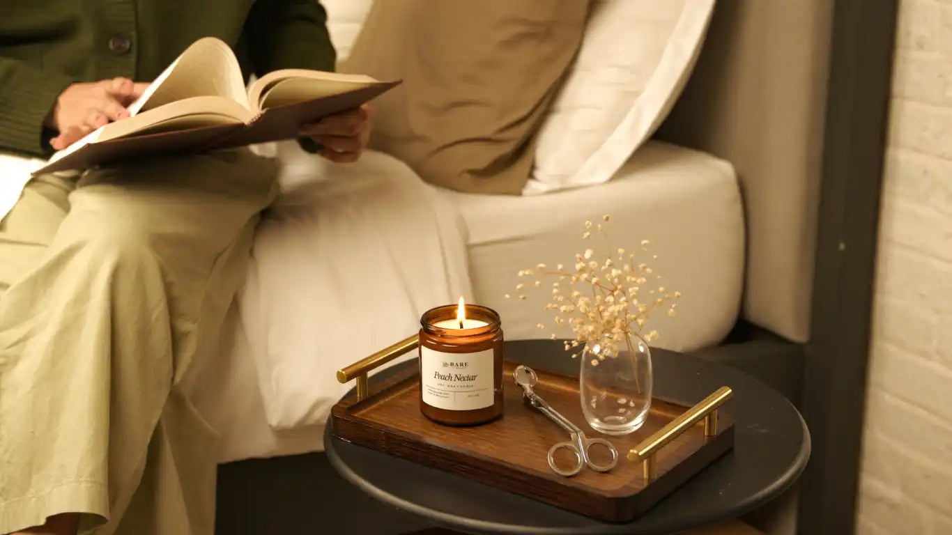 Person reading a book on their bed with a lit candle on their bedside placed on top of a wooden tray