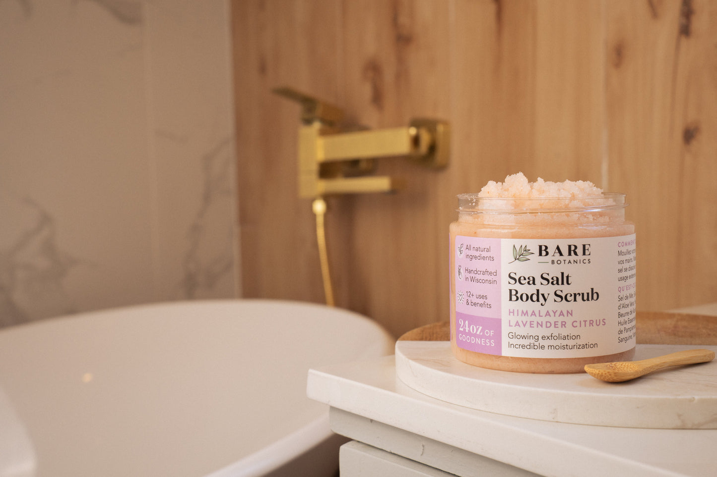 Jar of Bare Botanics Himalayan Sea Salt Body Scrub on a bathroom counter with a bathtub and wooden wall in the background.