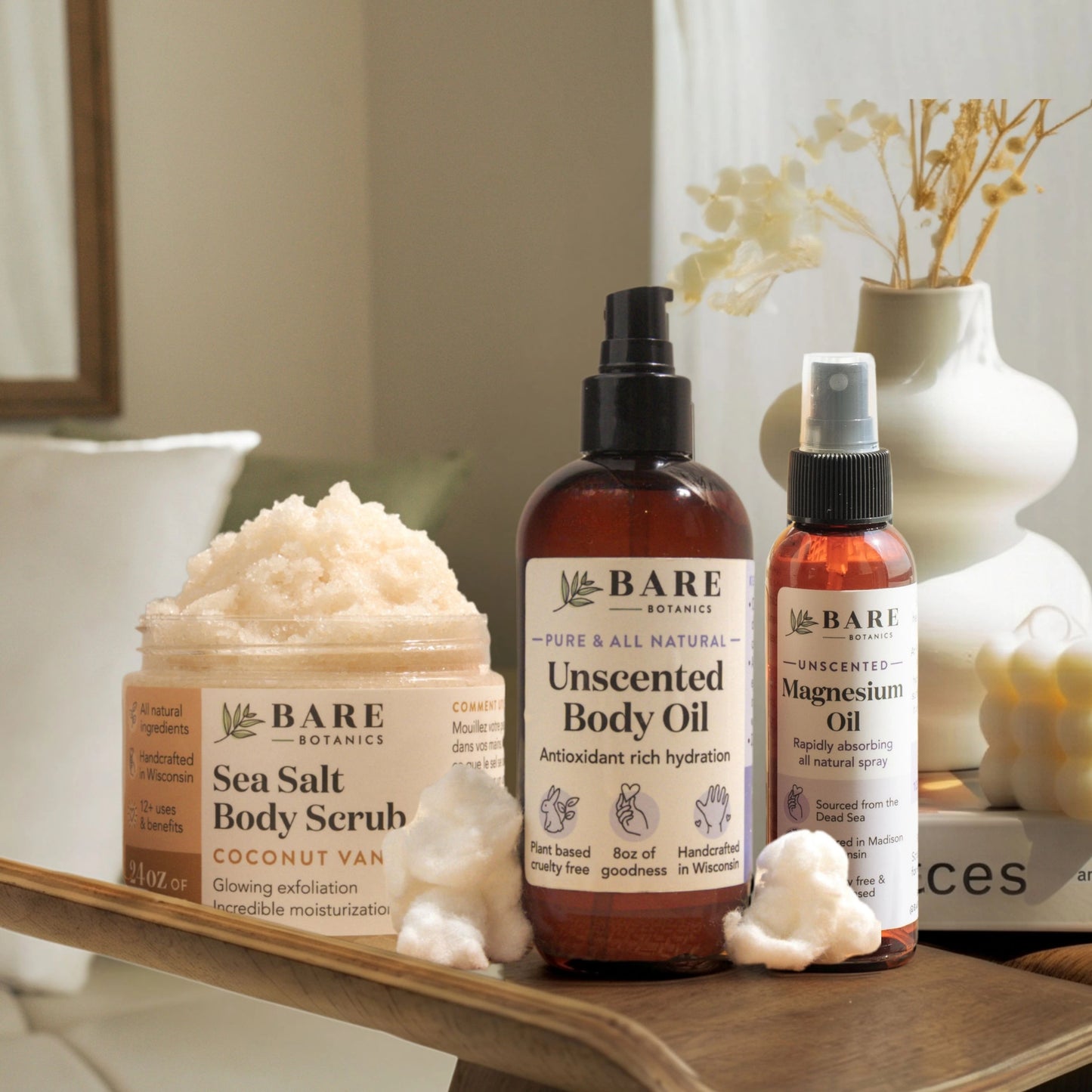 Skincare products from Bare Botanics with sea salt body scrub, unscented oil, and magnesium oil on a wooden platform with a vase and neutral background