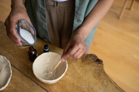 person mixing salts to create a scrub on a mixing bowl with bottles on a wooden table